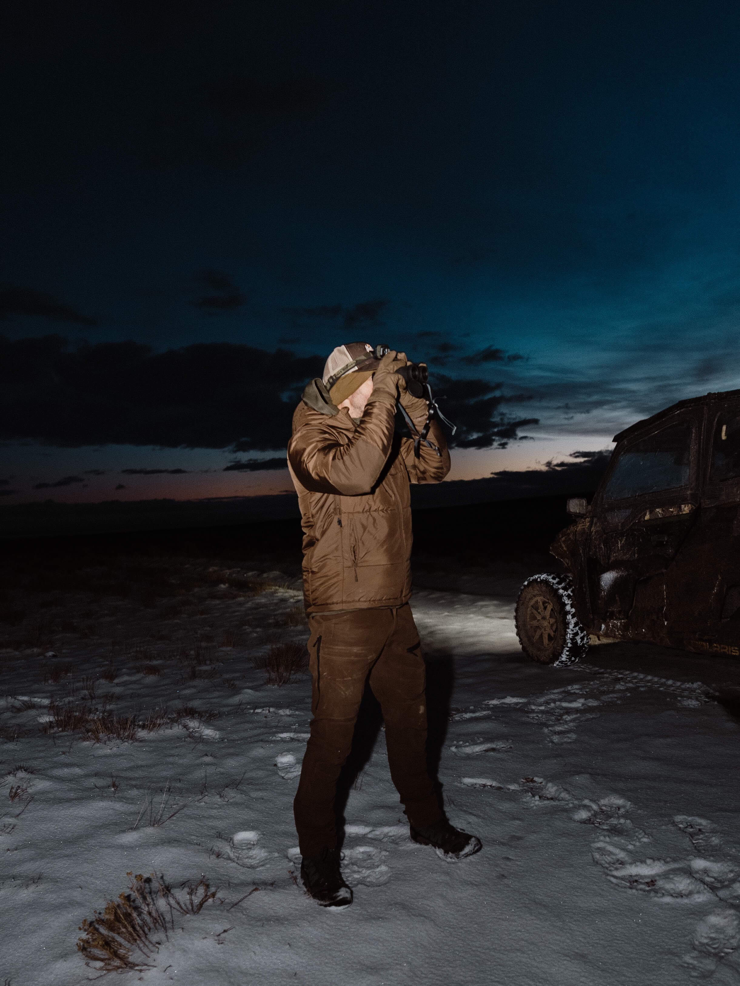 Man with binoculars beside an off-road vehicle at dusk